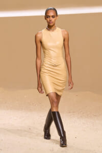 Model walks the runway in a form-fitting beige sleeveless dress with a thin belt and knee-high brown boots.