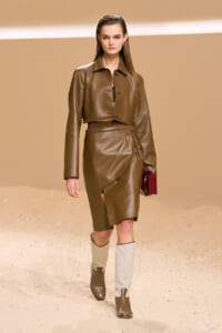 Model walks the runway in a brown cropped leather jacket with a matching wrap skirt, belt, and two-tone beige boots, carrying a burgundy clutch on a beige backdrop.