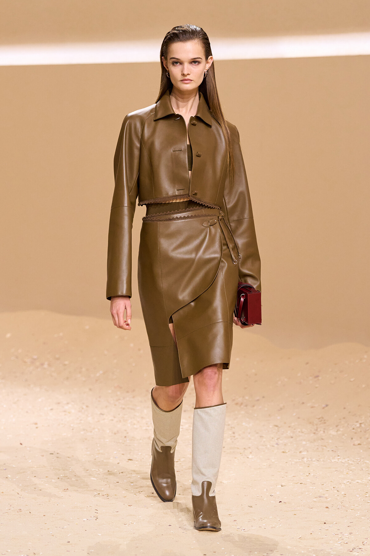 Model walks the runway in a brown cropped leather jacket with a matching wrap skirt, belt, and two-tone beige boots, carrying a burgundy clutch on a beige backdrop.