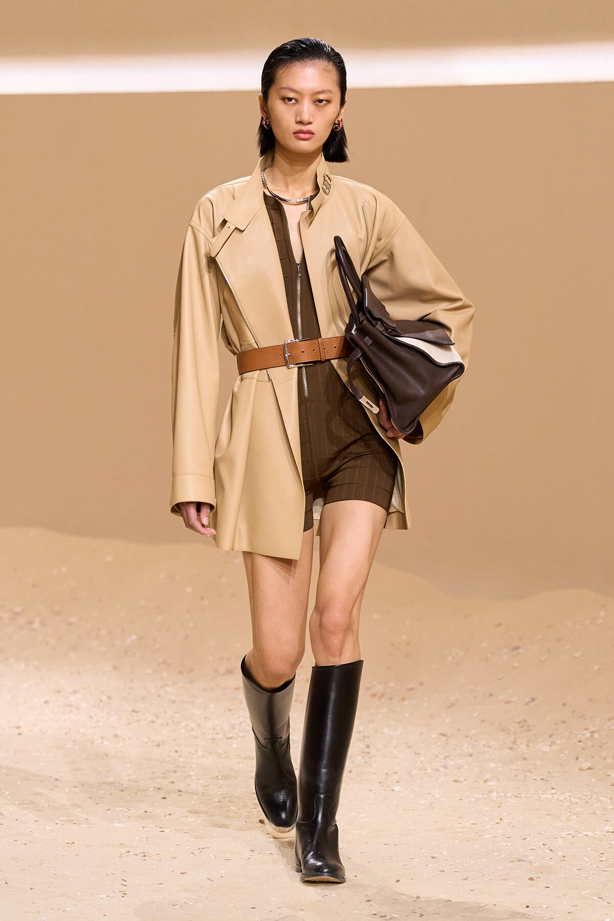Model on a runway wearing a tan trench coat cinched with a brown belt over a dark brown bodysuit, paired with black knee-high boots and a black handbag.