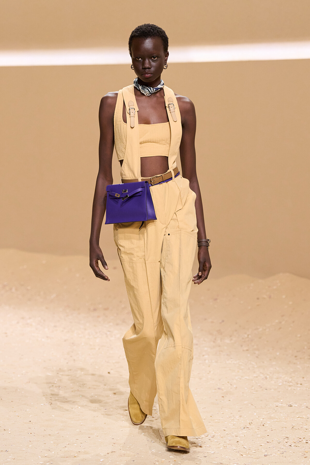 Female model walks a runway in a pale-yellow utility jumpsuit with a matching crop top, accessorized with a blue belt bag and scarf at the neck.