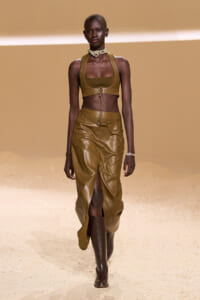 Model on a runway wearing a camel leather halter crop top and matching high-slit skirt with boots, accessorized with a choker and bracelet.