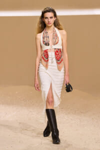 Model walks a beige runway in a white halter top and ribbed skirt, accessorized with a patterned scarf scarf and black knee-high boots.