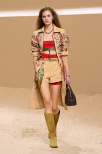 Model walks runway in a beige printed trench over a red bandeau, tan shorts with a belt, knee-high boots, and a black handbag on a sandy backdrop.