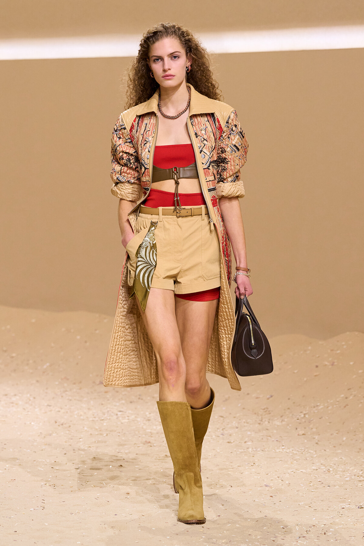 Model walks runway in a beige printed trench over a red bandeau, tan shorts with a belt, knee-high boots, and a black handbag on a sandy backdrop.