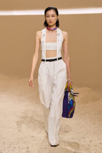 Model on a runway wearing white utility overalls with a bandeau top, black belt, and a colorful scarf at the neck, holding a multicolored bag.