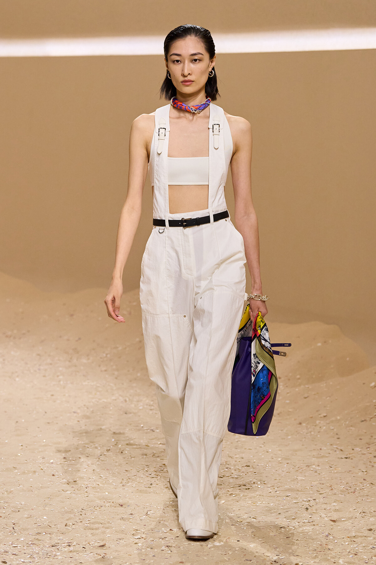 Model on a runway wearing white utility overalls with a bandeau top, black belt, and a colorful scarf at the neck, holding a multicolored bag.