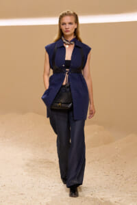 Model walking on a sandy runway in a navy sleeveless shirt-dress with a black harness belt, black top beneath, and a small black bag at her waist. A scarf and pendant necklace complete the look.