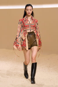 Model walks a runway in a red patterned kimono-style top over olive shorts and tall black boots, desert backdrop.