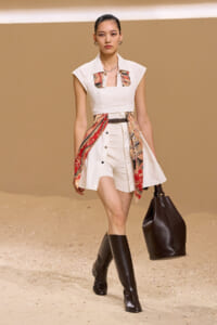 Model on a runway wearing a white sleeveless dress with colorful patterned scarf panels, a brown belt, black knee-high boots, and a large black handbag.
