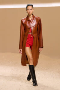 Model walks a runway in a brown leather long coat over a red outfit with knee-high black boots.