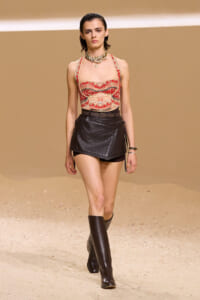 Model walking a runway in a desert-toned set, wearing a red patterned corset-style top, a black asymmetric leather mini skirt, tall black boots, and layered necklaces.