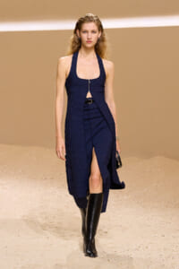 Model walking a runway in a navy ribbed halter dress with a front zipper and wrap detail, paired with black knee-high boots and a small clutch in hand.