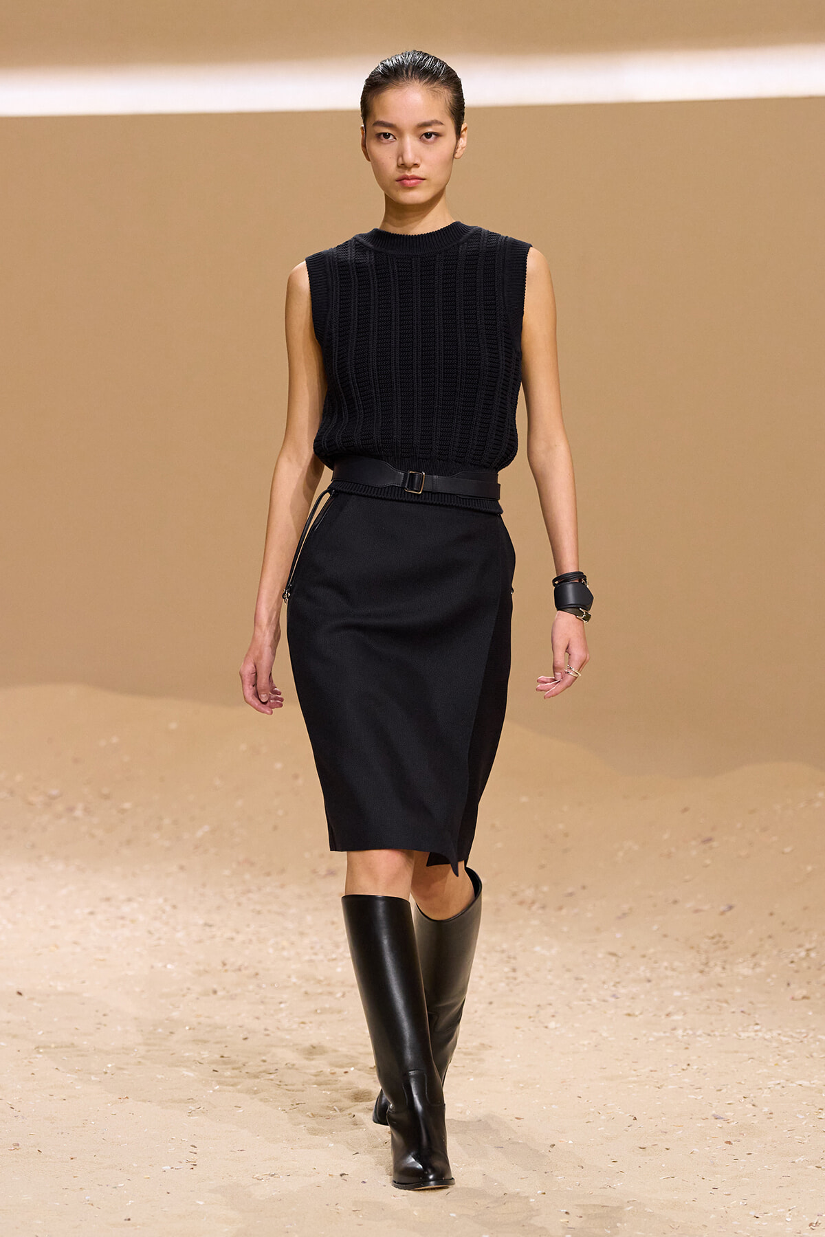 Female model on a fashion runway wearing a sleeveless black knit top, black pencil skirt, belt, and knee-high black boots.
