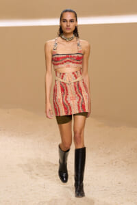 Model on a fashion runway wearing a red-and-beige patterned crop top and matching mini skirt with knee-high black boots .