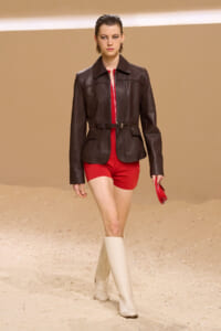 Model walking a sandy runway in a dark brown leather jacket with a belt, bright red shorts, and cream knee-high boots.