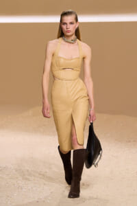 Model walking a runway in a tan quilted two-piece outfit with a cropped top, high-waisted skirt and knee-high brown boots, carrying a black bag.