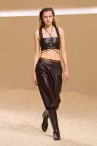 Model walks the runway in a black leather crop top with lace-up front, matching wide leather pants, brown belt, and knee-high boots on a sandy set.