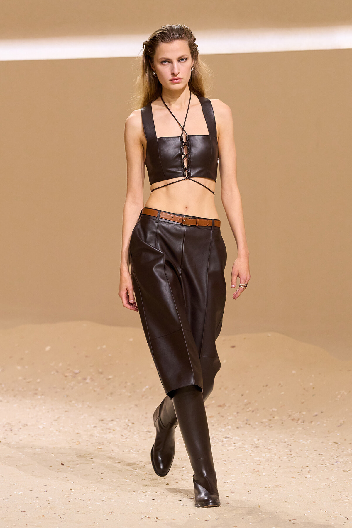 Model walks the runway in a black leather crop top with lace-up front, matching wide leather pants, brown belt, and knee-high boots on a sandy set.