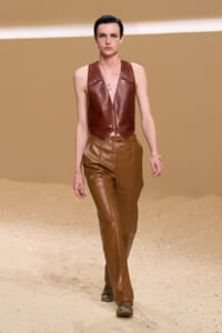 Model walks a sandy runway wearing a brown leather zip-front vest and matching high-waisted leather pants, accessorized with bracelets and a necklace.