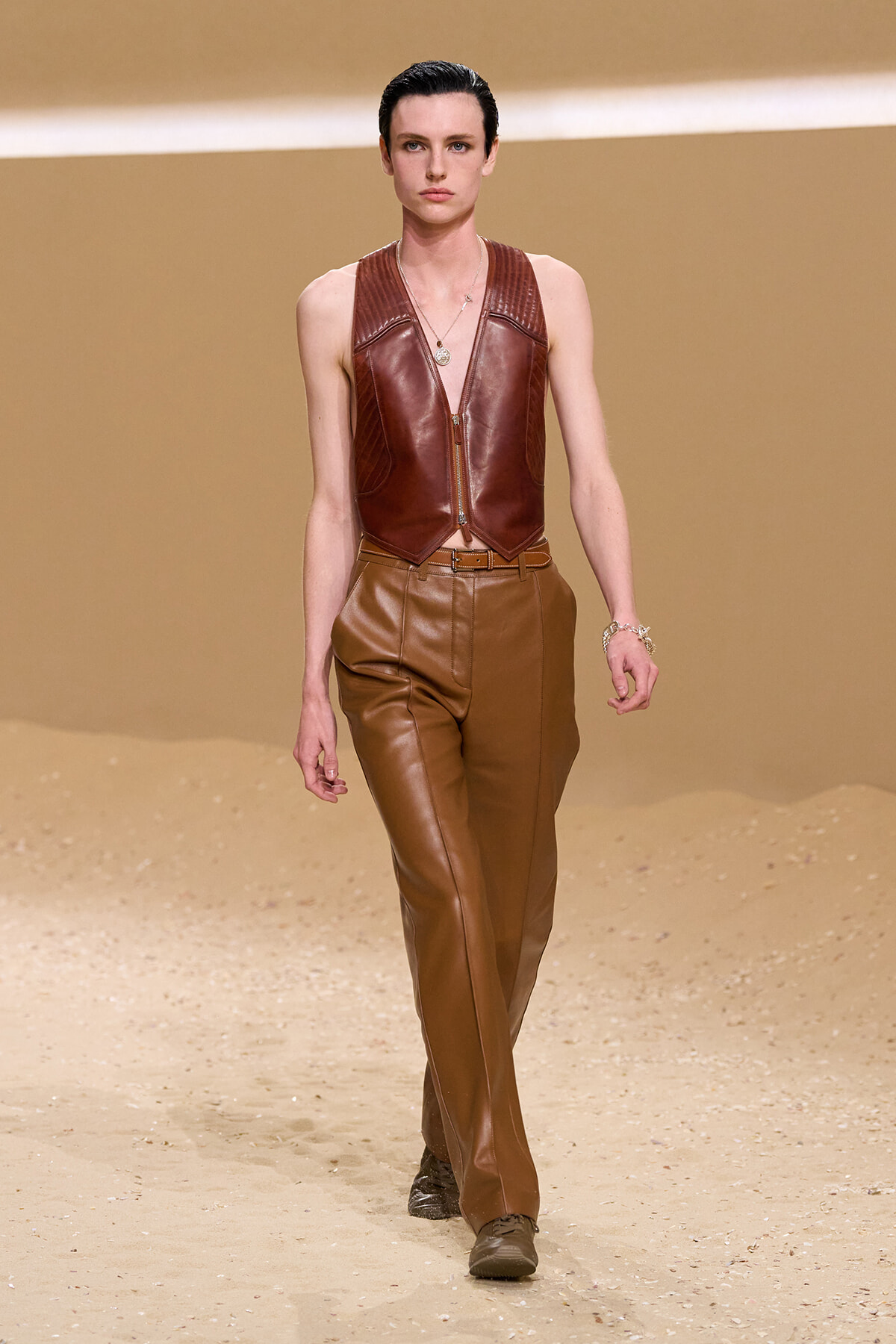 Model walks a sandy runway wearing a brown leather zip-front vest and matching high-waisted leather pants, accessorized with bracelets and a necklace.