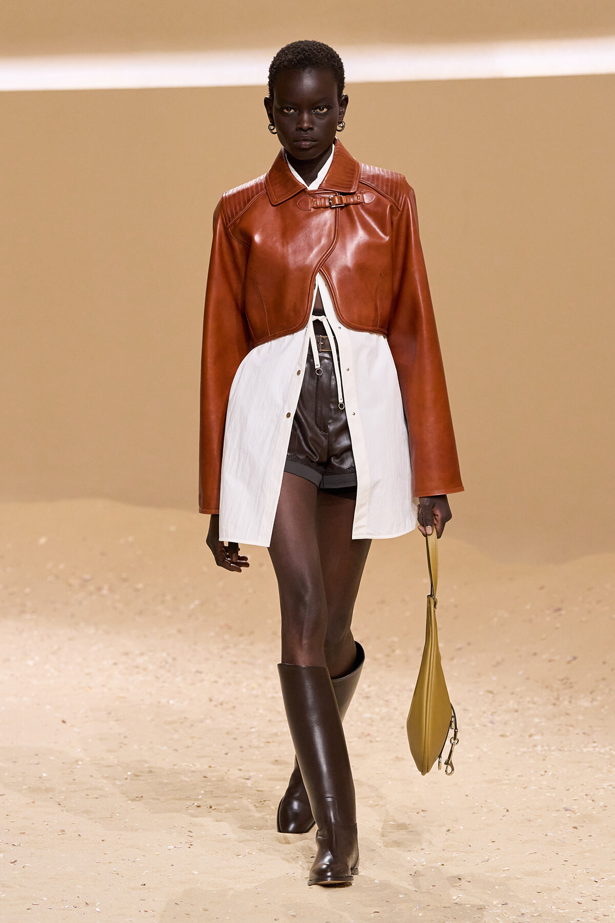 Model walks a runway in a rust-brown cropped leather jacket over a white layered dress with black shorts and tall brown boots, carrying a mustard-yellow handbag.