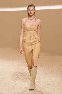 Model walks the runway in a tan coordinated outfit: a quilted crop top with a front zipper and matching high-waisted pencil skirt with front zip and pockets, paired with knee-high boots.
