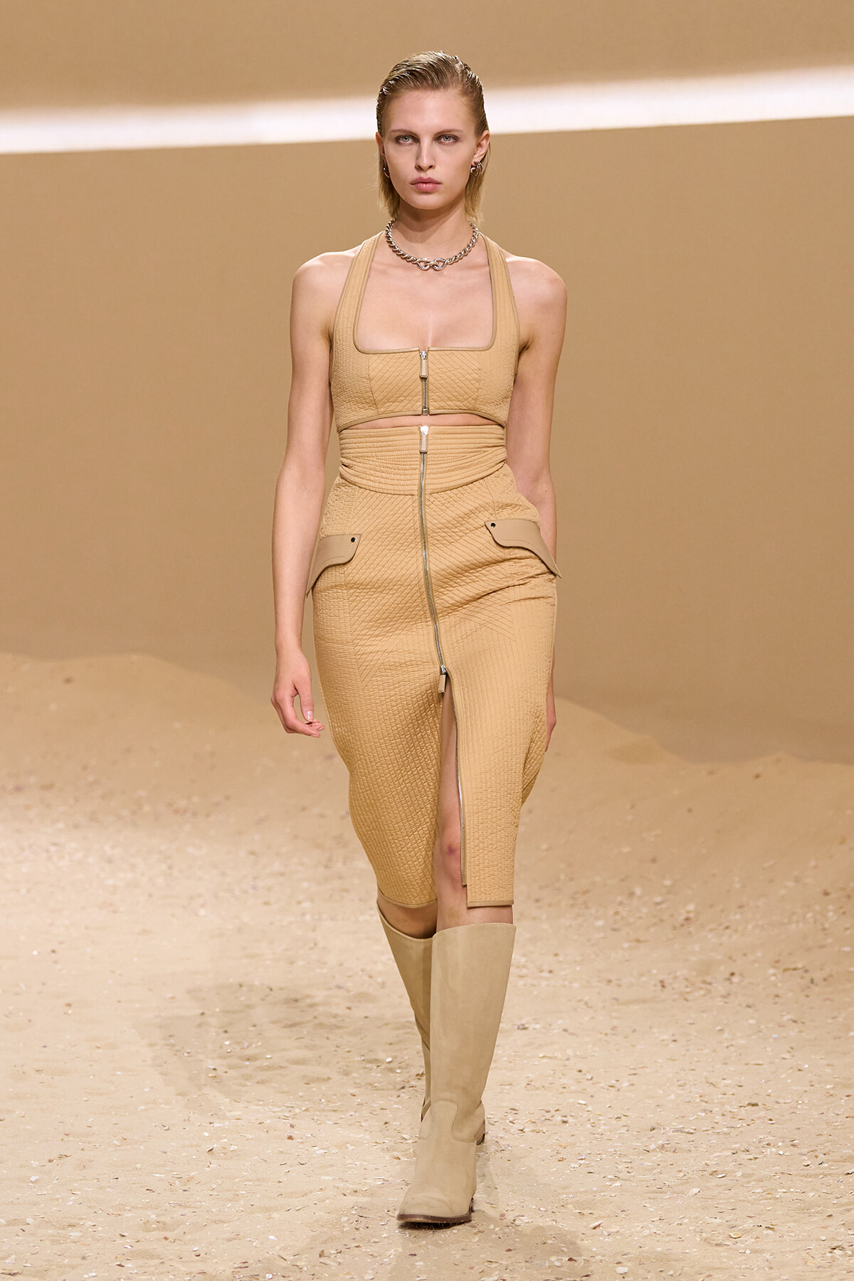 Model walks the runway in a tan coordinated outfit: a quilted crop top with a front zipper and matching high-waisted pencil skirt with front zip and pockets, paired with knee-high boots.