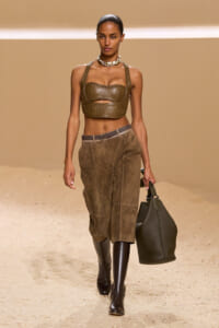 Runway model in an earthy desert-toned setting wearing a brown leather strapless crop top, suede pants, tall boots, and a large dark tote bag.