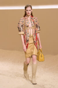 Model walking in a beige-toned runway outfit: patterned kimono-style jacket over a pale pink top, tan skirt, knee-high boots, and a mustard handbag.