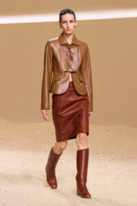 Model on a runway wearing a tan cropped leather jacket with lace-up front, a rust brown leather pencil skirt, and matching knee-high boots, against a sandy backdrop.