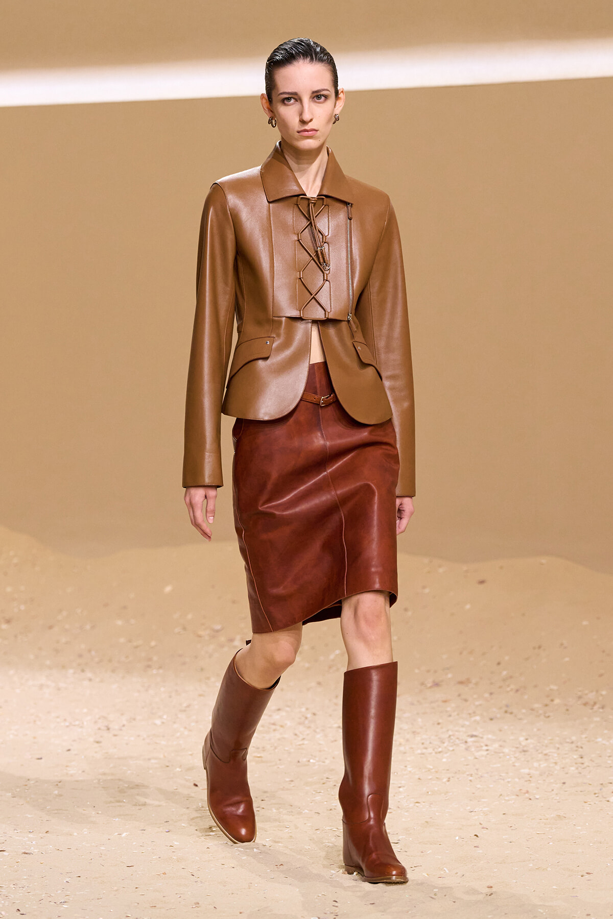 Model on a runway wearing a tan cropped leather jacket with lace-up front, a rust brown leather pencil skirt, and matching knee-high boots, against a sandy backdrop.