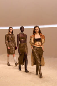 Three models walk a runway in brown and olive leather outfits against a tan backdrop, earth-toned and cohesive in style.