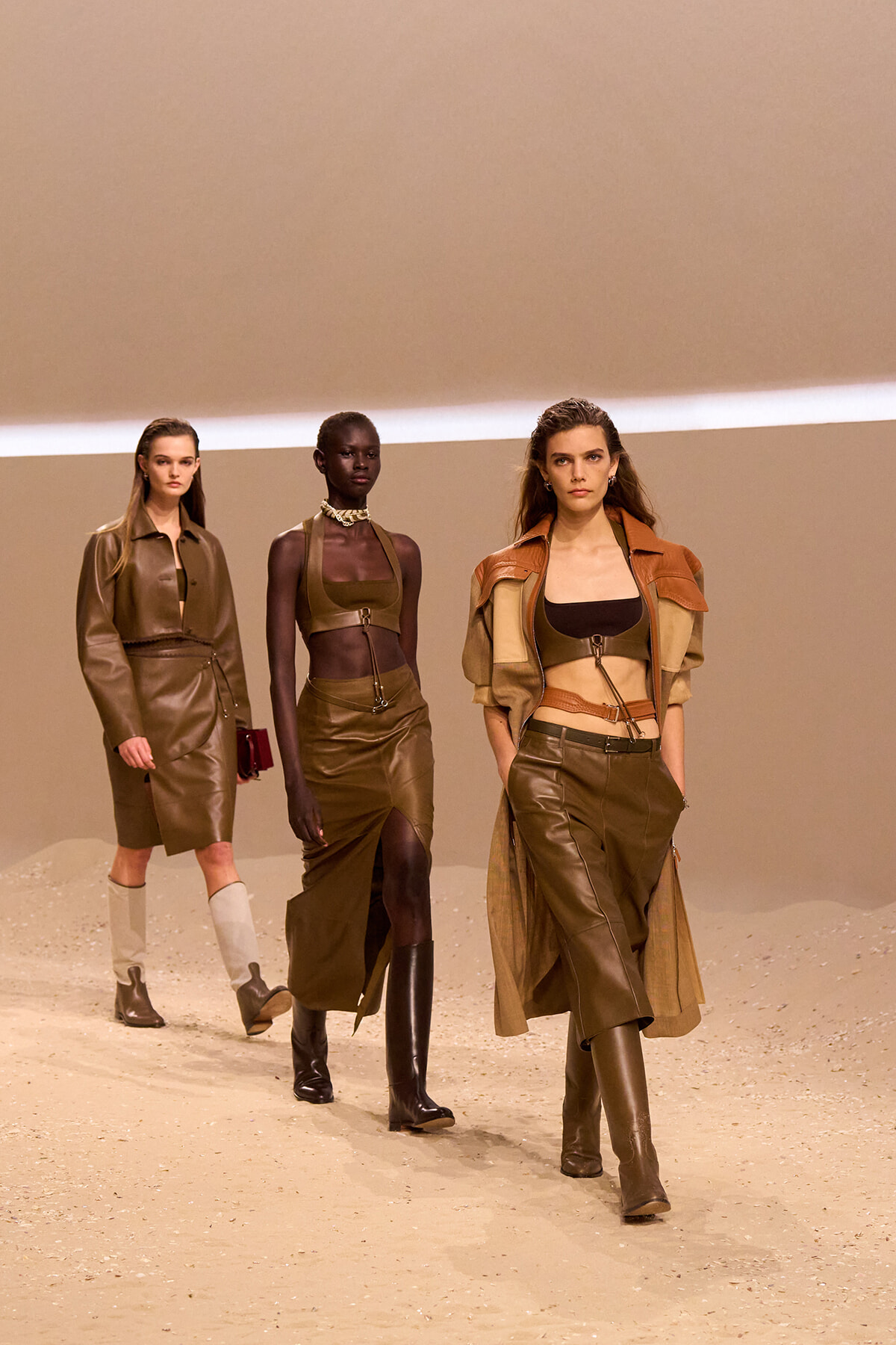 Three models walk a runway in brown and olive leather outfits against a tan backdrop, earth-toned and cohesive in style.