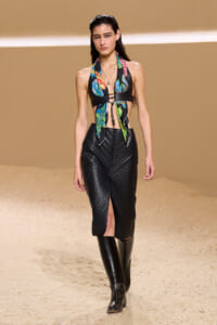 Model walking a runway in a colorful tropical halter top and black quilted midi skirt with knee-high boots on a beige catwalk surface.