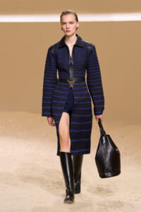 Model on a runway wearing a navy striped quilted coat with leather shoulders, a gold chain belt, knee-high boots, and a black handbag.