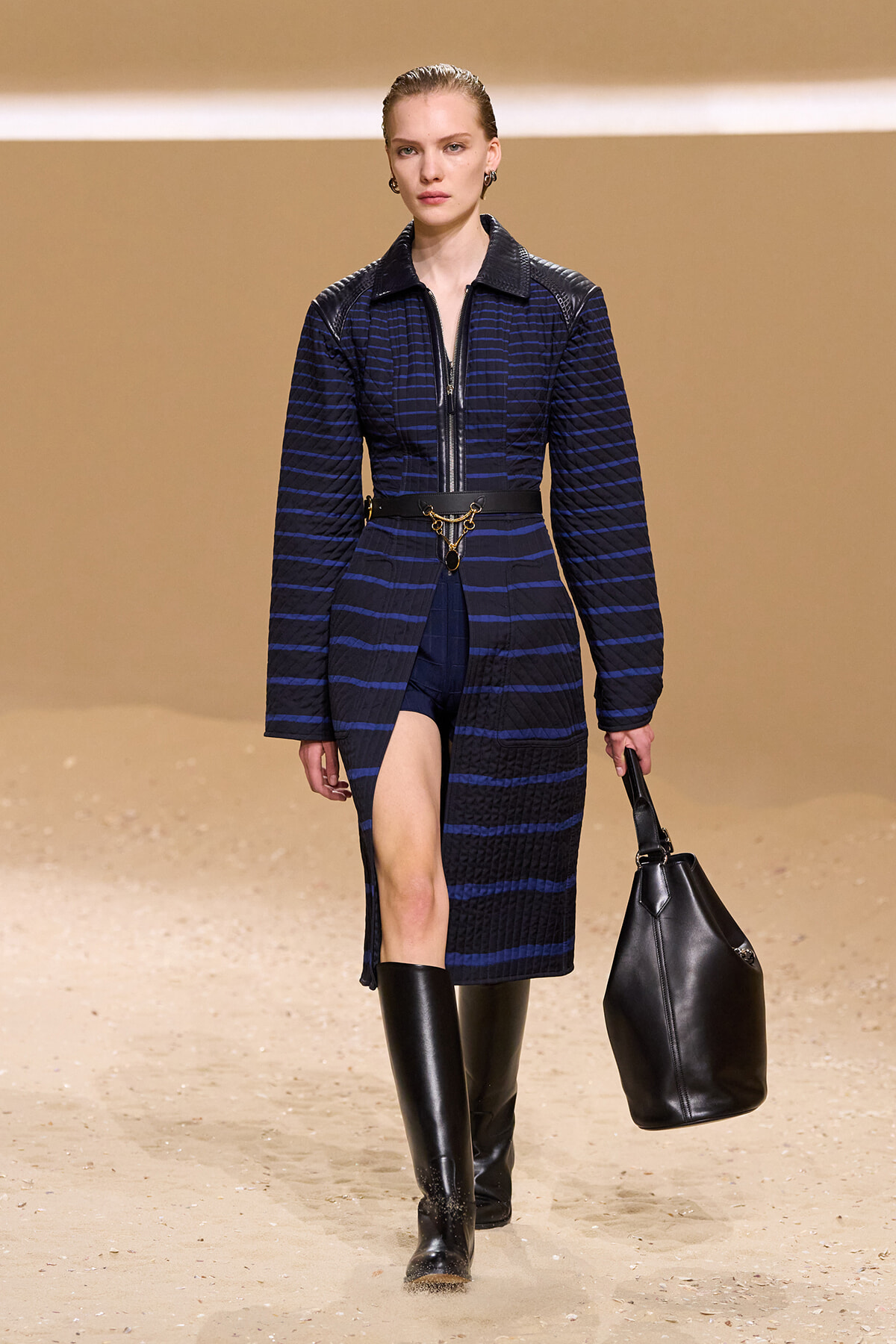 Model on a runway wearing a navy striped quilted coat with leather shoulders, a gold chain belt, knee-high boots, and a black handbag.