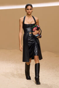 Model walking a runway in a black leather corset top, long matching vest, and knee-high boots, clutching a colorful scarf.