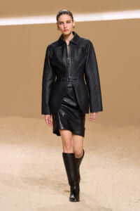 Model walking the runway in a black quilted leather jacket with a belt, paired with a black mini skirt and knee-high boots.