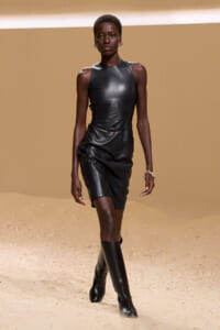 Model walking on a sandy runway in a black sleeveless leather top, belt, and knee-high boots.