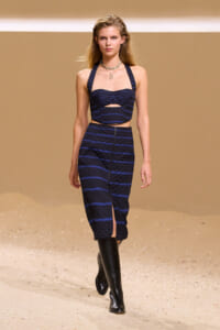 Runway model wearing a navy striped quilted crop top and matching high-waisted midi skirt with a thigh slit, black knee-high boots, beige backdrop.