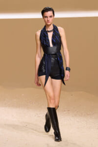 Model walking the runway in a black leather romper with a wide belt and a blue striped scarf, paired with knee-high black boots, on a sandy backdrop.