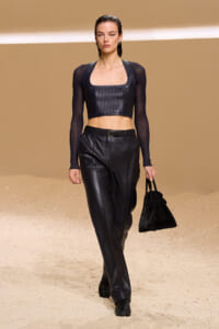 Runway model in a black leather crop top and matching wide-leg pants, carrying a black handbag.