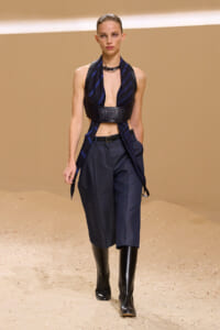 Runway model wearing a navy striped halter crop top with a wide black leather band, a matching scarf, high-waisted navy trousers, and black knee-high boots.