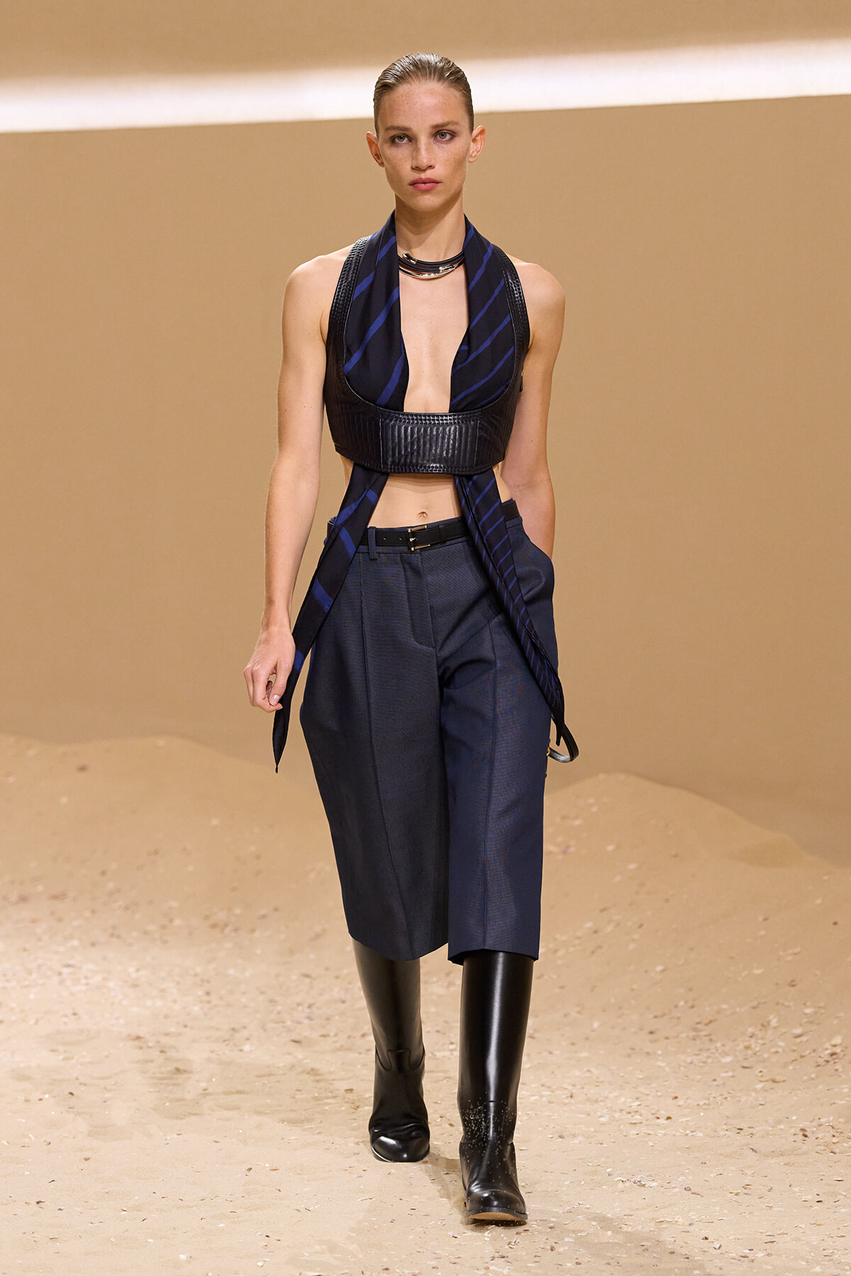 Runway model wearing a navy striped halter crop top with a wide black leather band, a matching scarf, high-waisted navy trousers, and black knee-high boots.