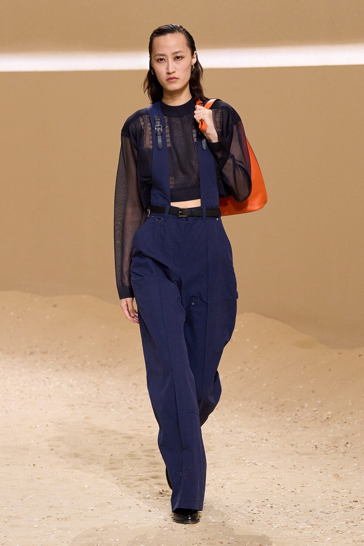 Runway model wearing navy utility pants and a sheer black top with harness straps, carrying an orange bag against a sandy beige backdrop.
