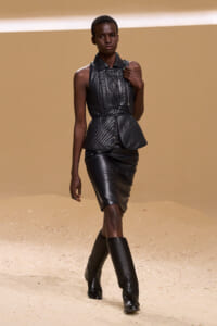 Model walks a runway in a black quilted leather sleeveless top with peplum waist, matching knee-length skirt, and tall boots.