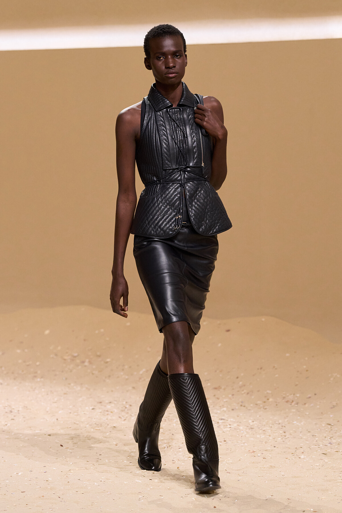 Model walks a runway in a black quilted leather sleeveless top with peplum waist, matching knee-length skirt, and tall boots.