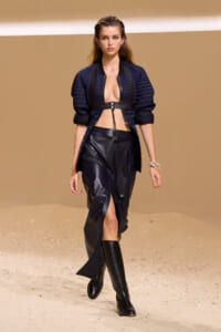 Model walking a runway in a navy striped jacket, black bralette harness, and leather midi skirt with a front slit, paired with tall black boots on a sandy backdrop.