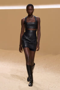 Model walking a fashion runway in a black quilted two-piece outfit (crop top and mini skirt) with knee-high boots on a sandy beige backdrop